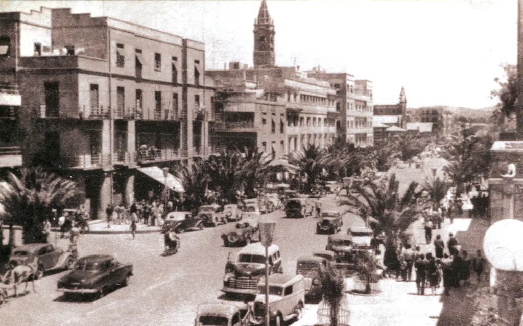 Harnet Avenue in Asmara in the 1930s, palm trees down the middle, cars and buses, and the cathedral tower behind