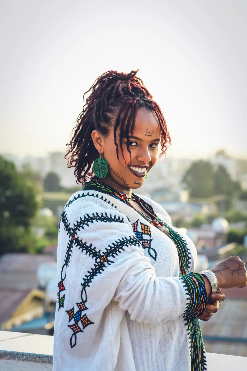 A smiling young woman in a white cotton dress with a bright embroidered border down the front, green beaded earrings and traditional facial markings, on a rooftop at golden hour