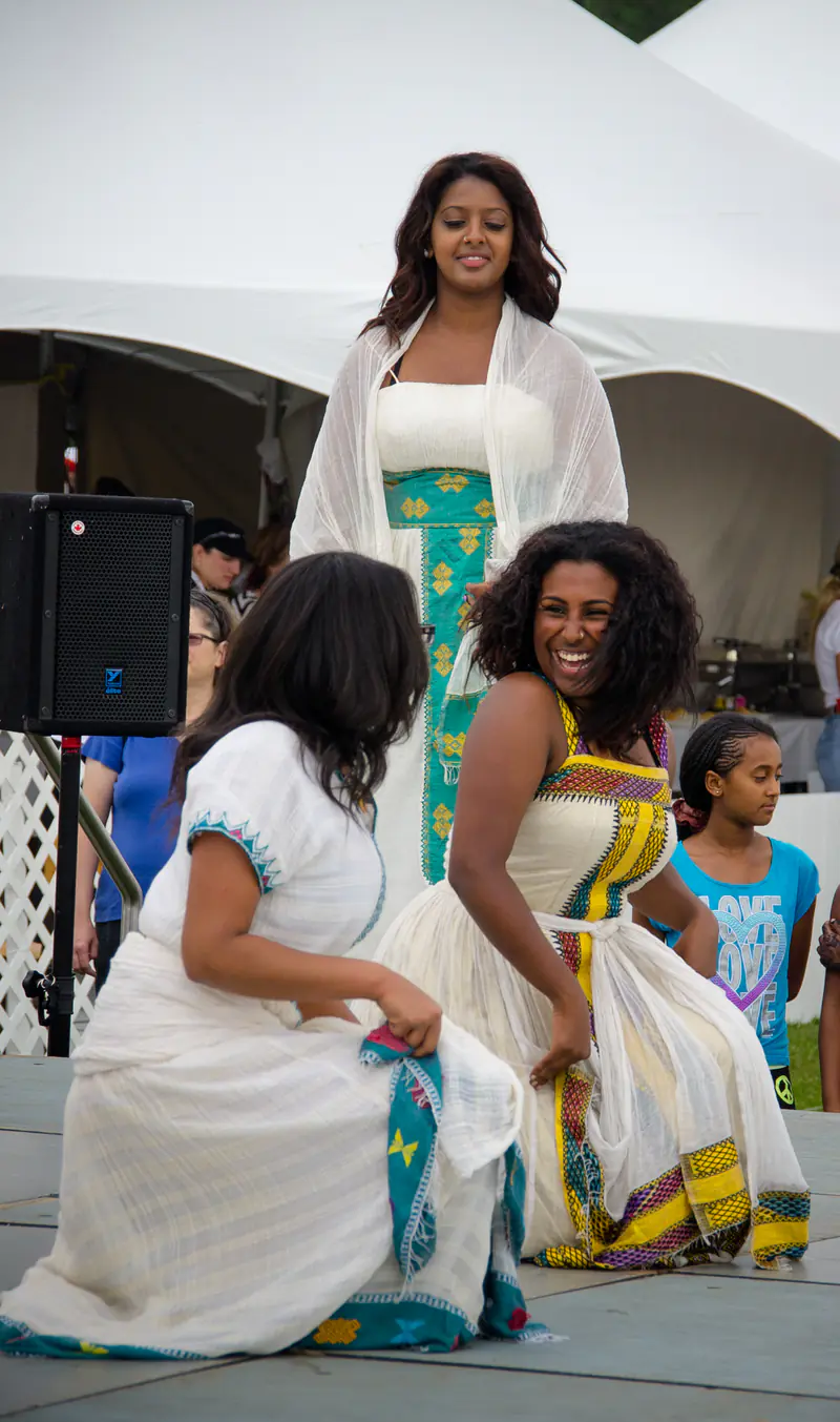 Three young women in white dresses with coloured borders dancing at an Eritrean community festival, one laughing