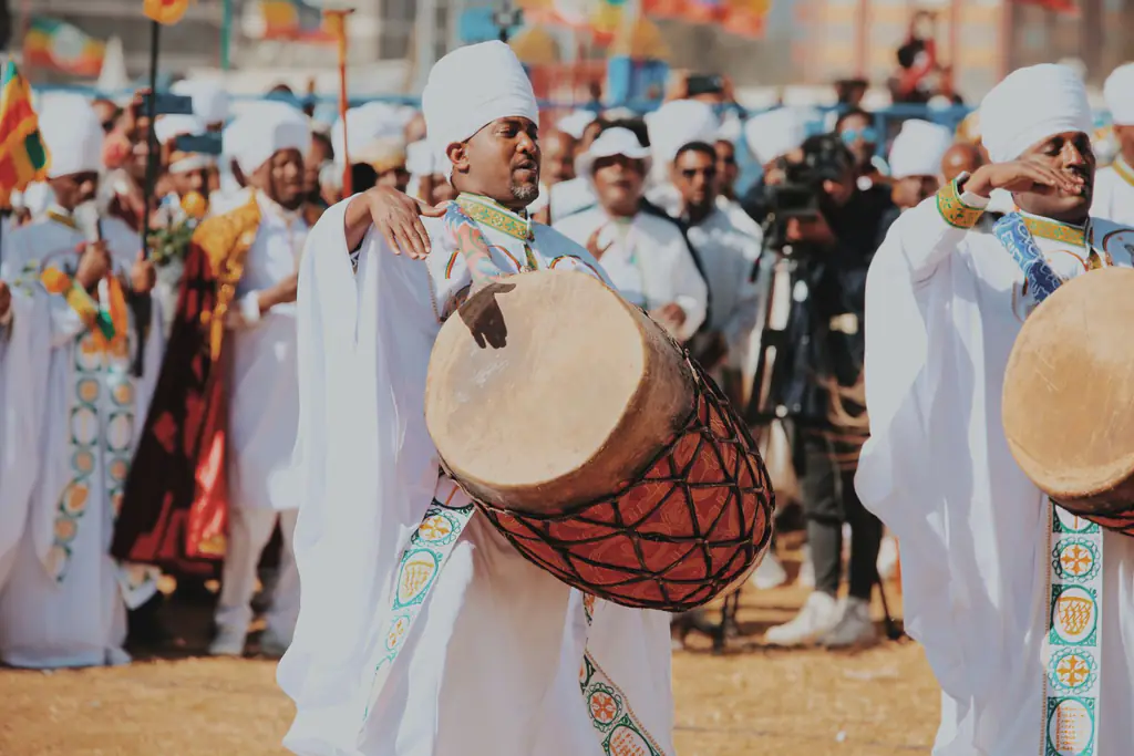 A drummer in white robes and a white turban playing a large kebero drum slung at his hip, in a crowd at a festival