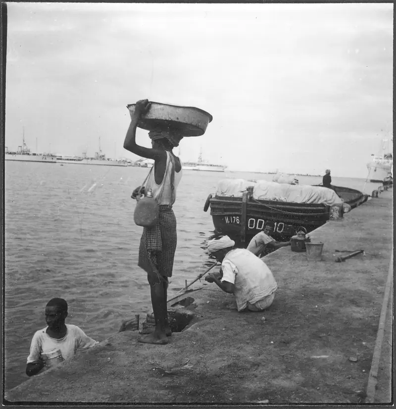 Porters on the quay at Massawa in 1939, one carrying a basin on his head, with a loaded boat and ships behind