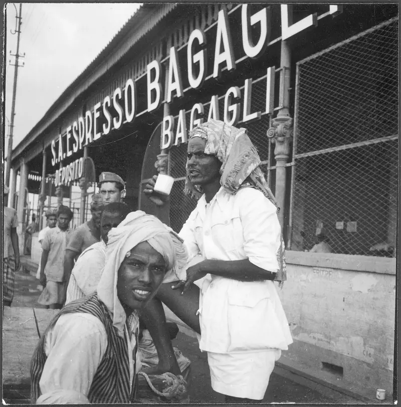 A tea seller in a turban lifting a small cup outside the baggage office on the Massawa waterfront in 1939, with a smiling customer in front