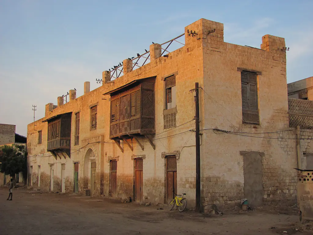 Carved wooden balconies on a coral-stone house in Massawa's old town, glowing in low evening sun, with a yellow bicycle leaning on the wall