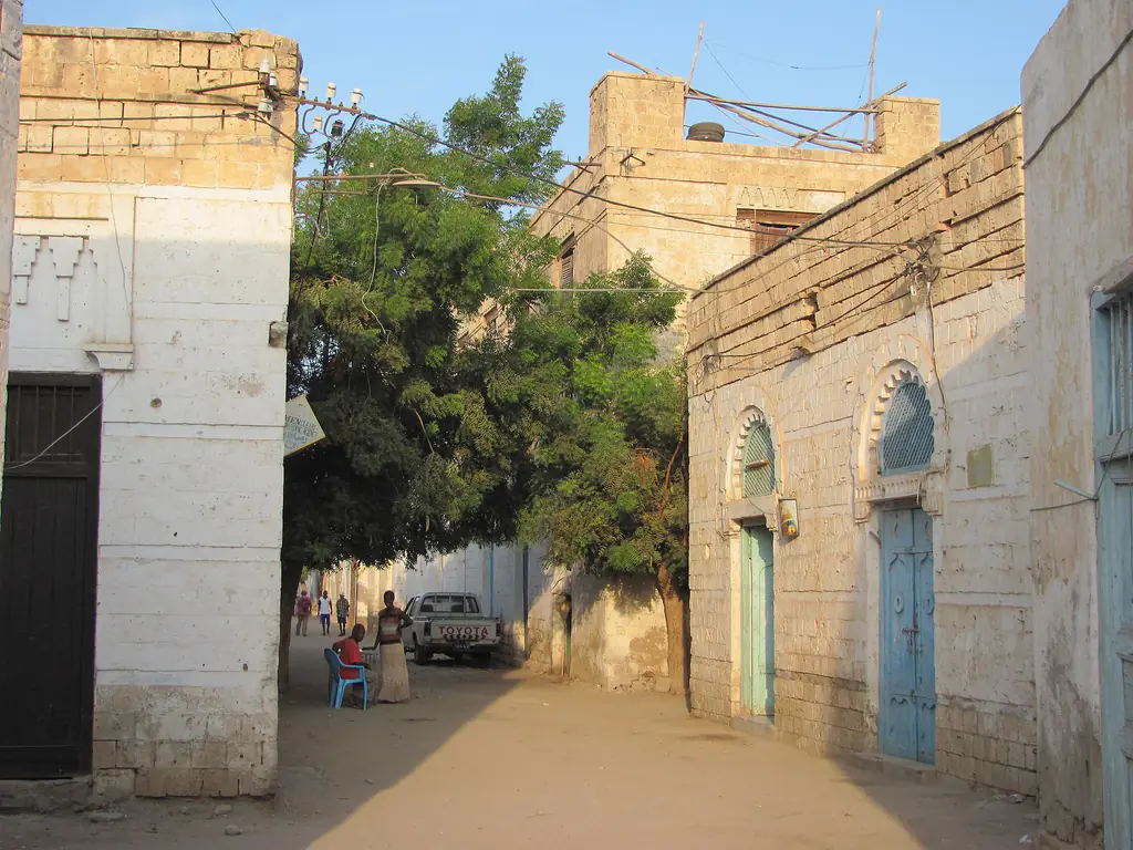 A sandy lane in Massawa's old town in evening light, with blue doors, a big tree and neighbours sitting outside
