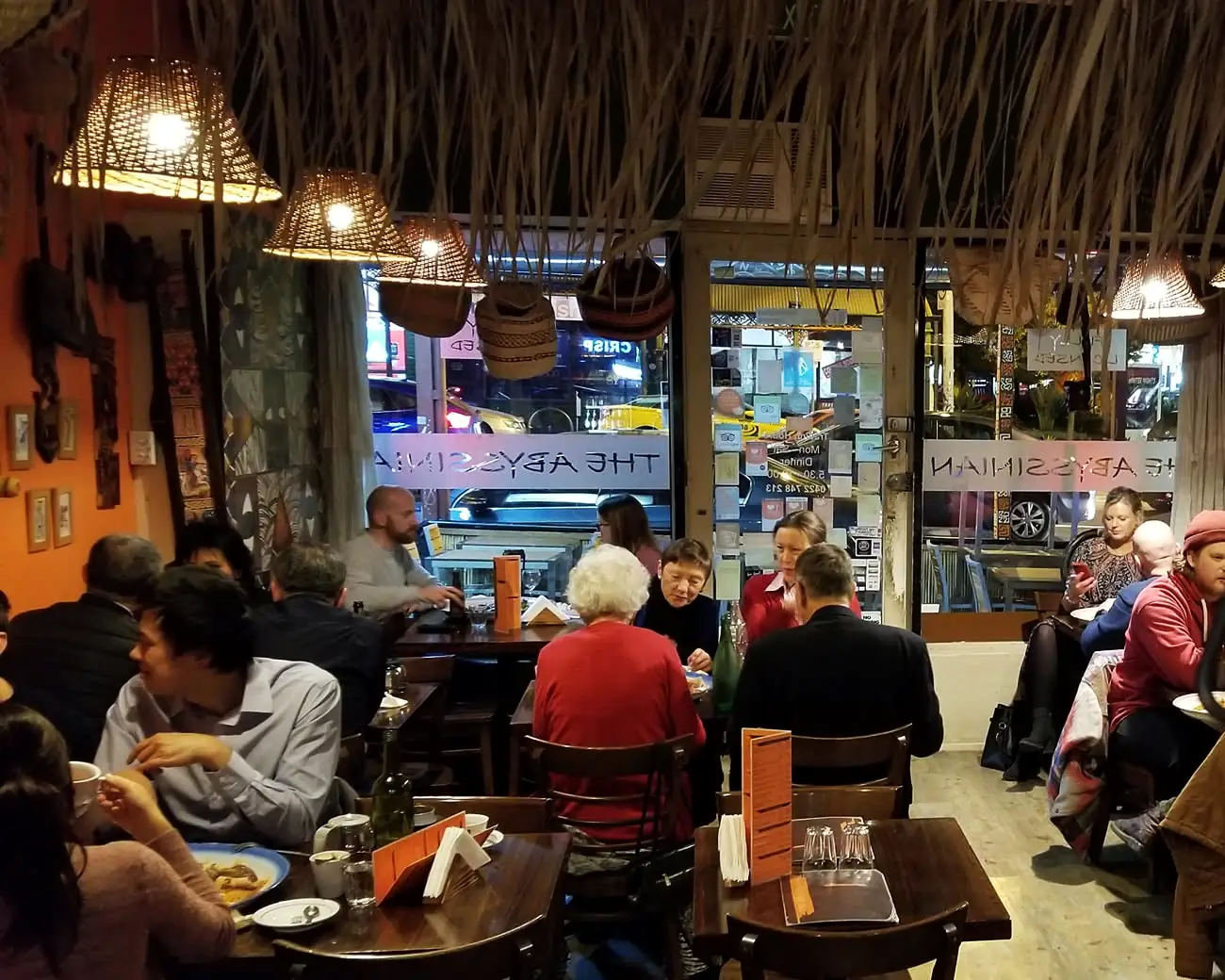 The dining room on a weeknight, guests at the tables under the thatch and the woven lanterns, the street through the window